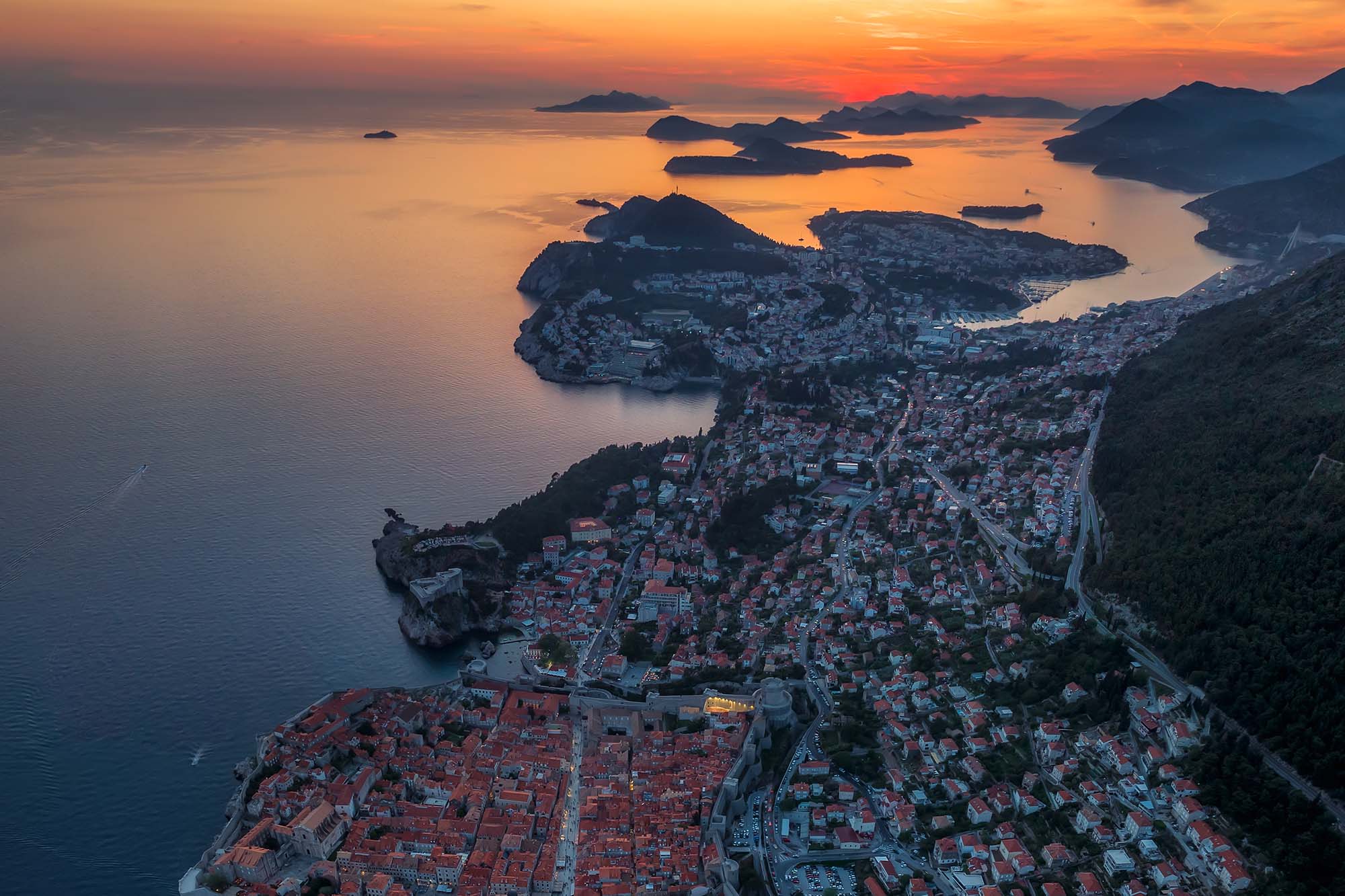 Amazing aerial panoramic view of the picturesque town of Dubrovnik with the old town, illuminated streets and buildings and marina with boats at fiery sunset.